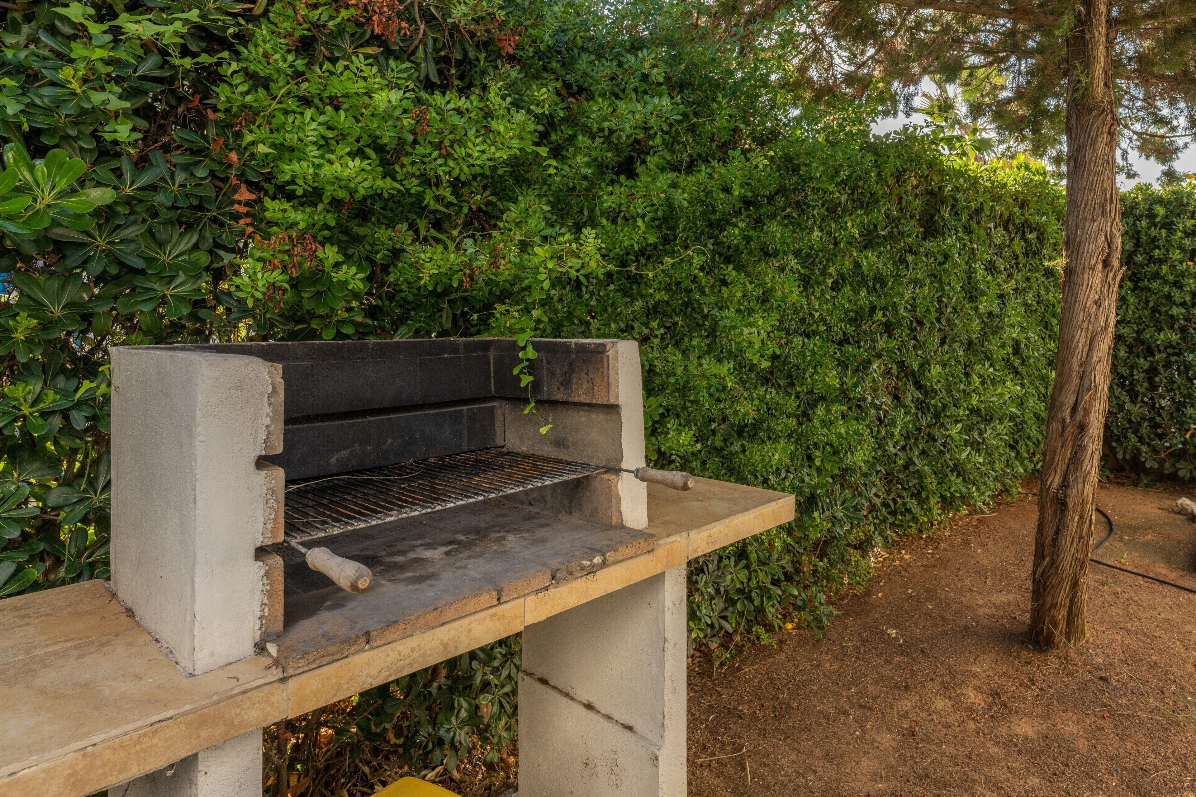 Un barbecue installé à l’extérieur, avec une base en béton et une grille en métal, entouré d’un feuillage verdoyant et dense.