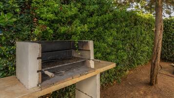 Un barbecue installé à l’extérieur, avec une base en béton et une grille en métal, entouré d’un feuillage verdoyant et dense.