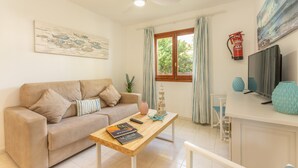 Ein Wohnzimmer mit einer Couch, einem Couchtisch aus Holz mit Büchern und einer Fernbedienung, einem Fernseher auf einem Standfuß, einem Feuerlöscher und einem Fenster mit Vorhängen.