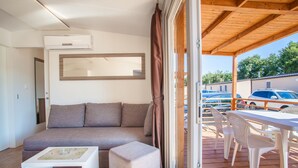 Ein modernes Wohnzimmer mit einer Couch, Hocker und einem kleinen Tisch. Ein großes Fenster mit Vorhängen sowie eine Holzterrasse mit Stühlen und einem Tisch sind ebenfalls zu sehen.