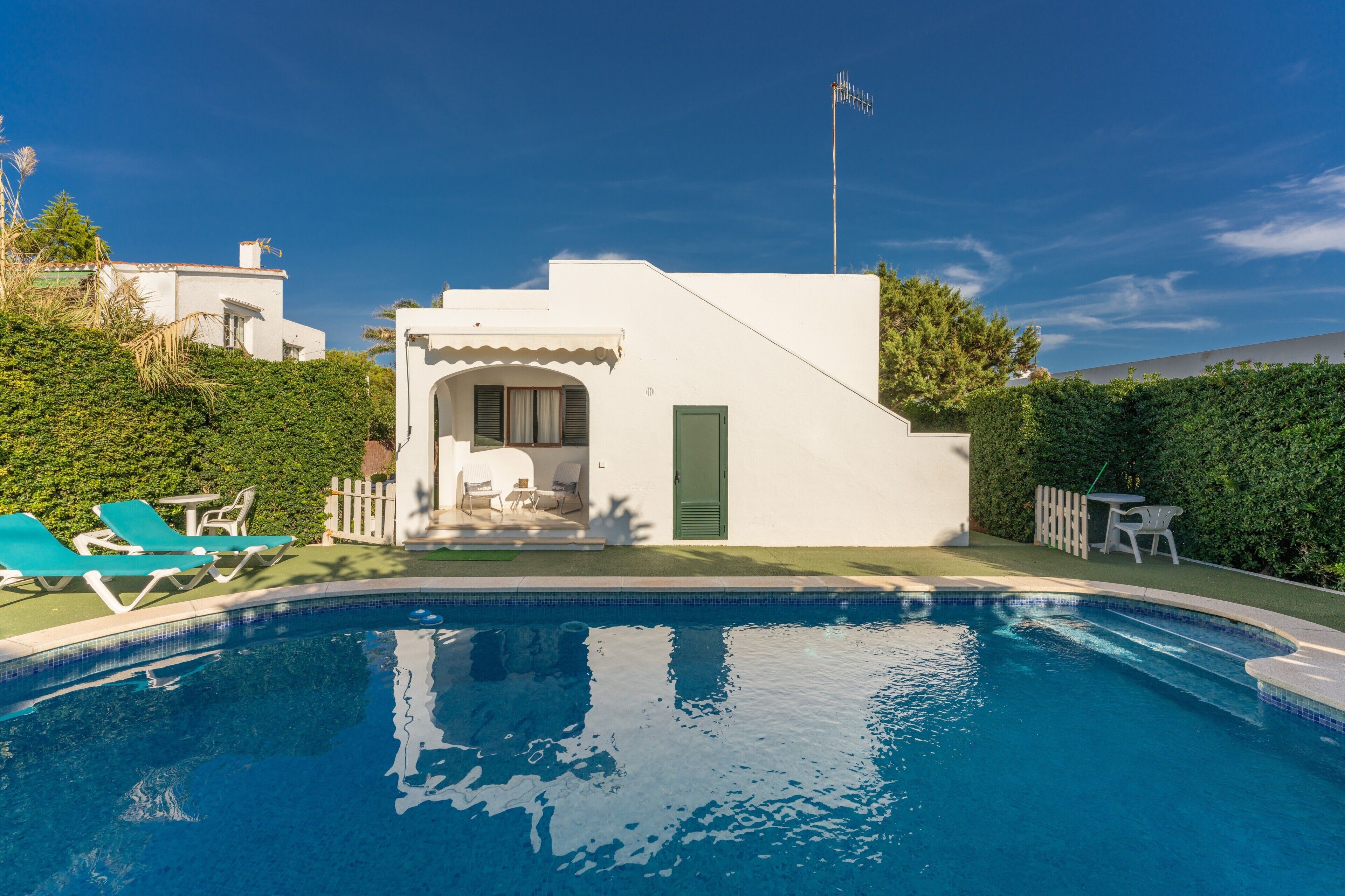 Una piscina con sillas de descanso, un pequeño patio con mesa y sillas, y un edificio blanco con una puerta y una ventana.