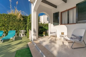 A covered outdoor seating area with lounge chairs and a table, a white wall with a window, and a hedge in the background.