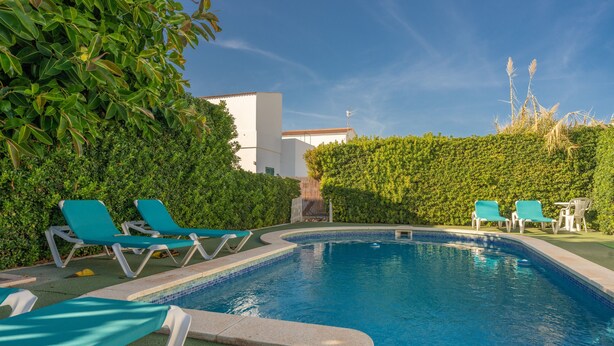 Una piscina con lettini, circondata da vegetazione e un edificio bianco.