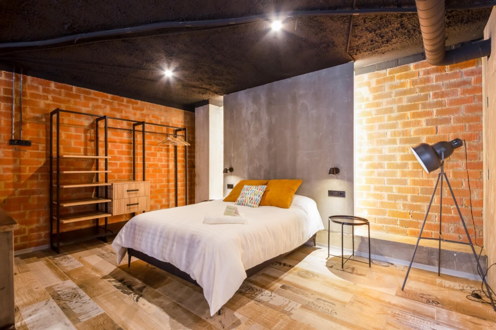 Un dormitorio con cama, suelo de madera, paredes de ladrillo, estante metálico y lámpara de pie.
