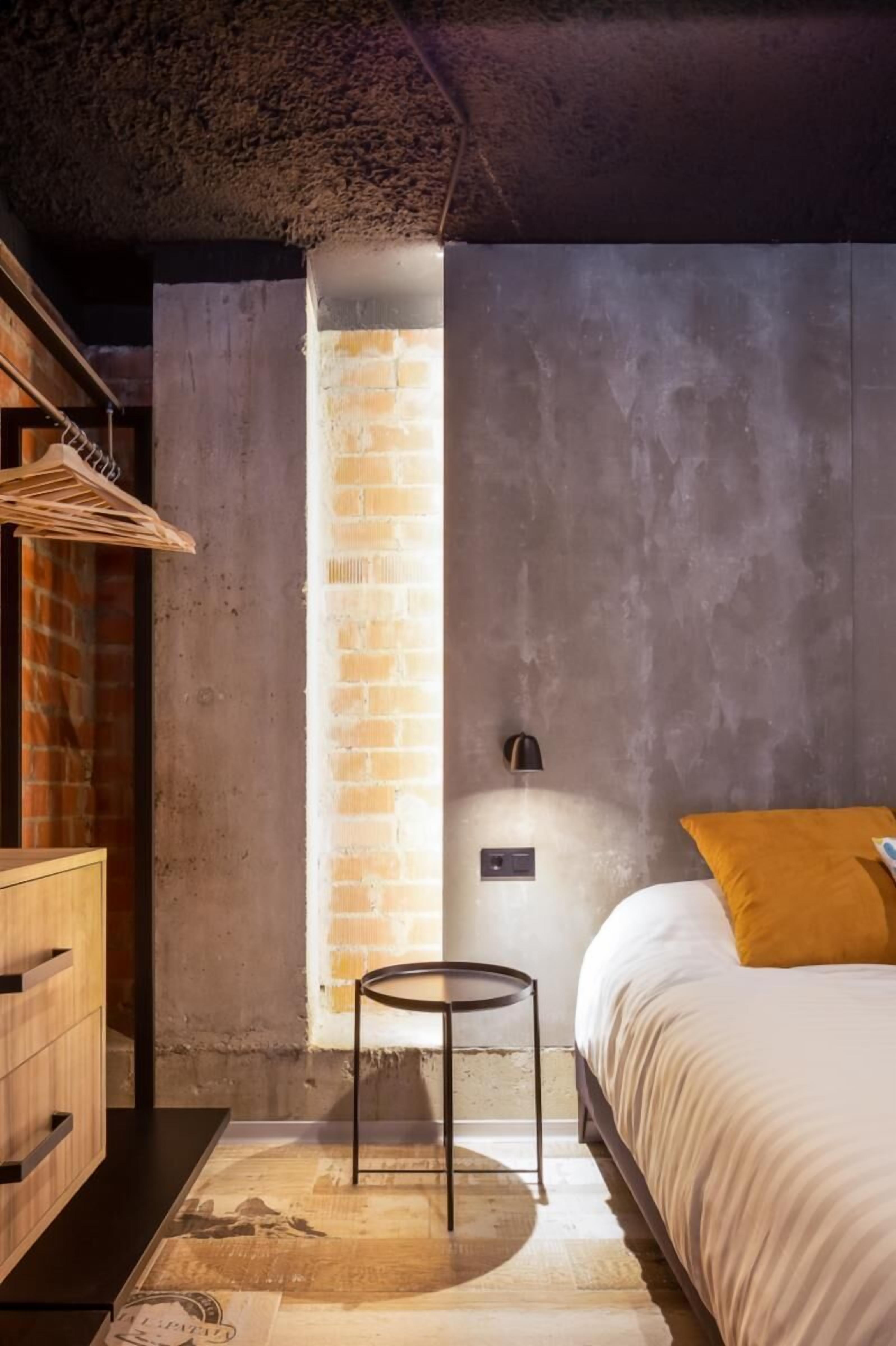Un dormitorio con pared de ladrillo, cómoda de madera, mesa redonda, cama con almohada amarilla y una lámpara.