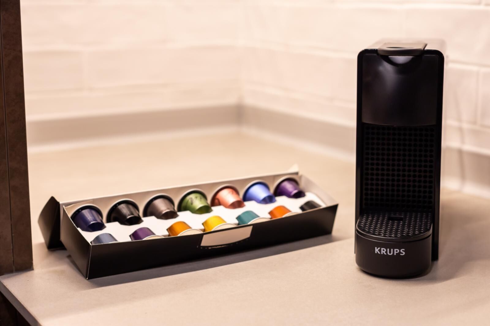 Una cafetera Krups negra junto a una caja de cápsulas de café de colores sobre una encimera.