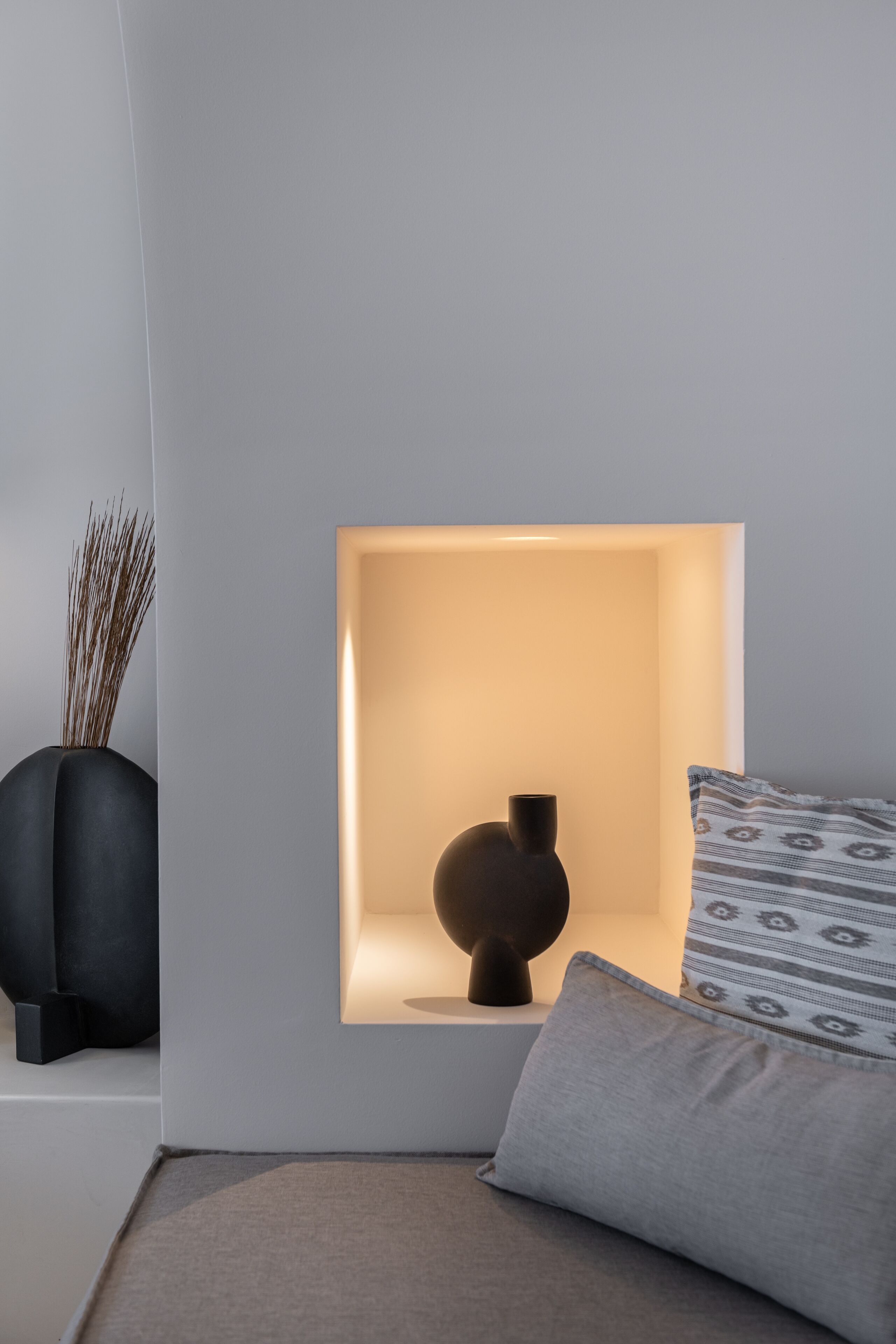 Una habitación moderna con diseño minimalista, que incluye una lámpara fijada a la pared, un jarrón oscuro con plantas secas y un cojín estampado sobre un sofá de tonos neutros.