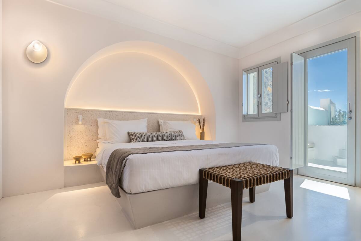 A modern bedroom with a curved ceiling, a large bed, bedside tables, a bench, and a view through a glass door.