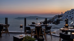 A dining area with a table set for two, overlooking a coastal town with white buildings. - Akra Suites & Spa (Santorini)