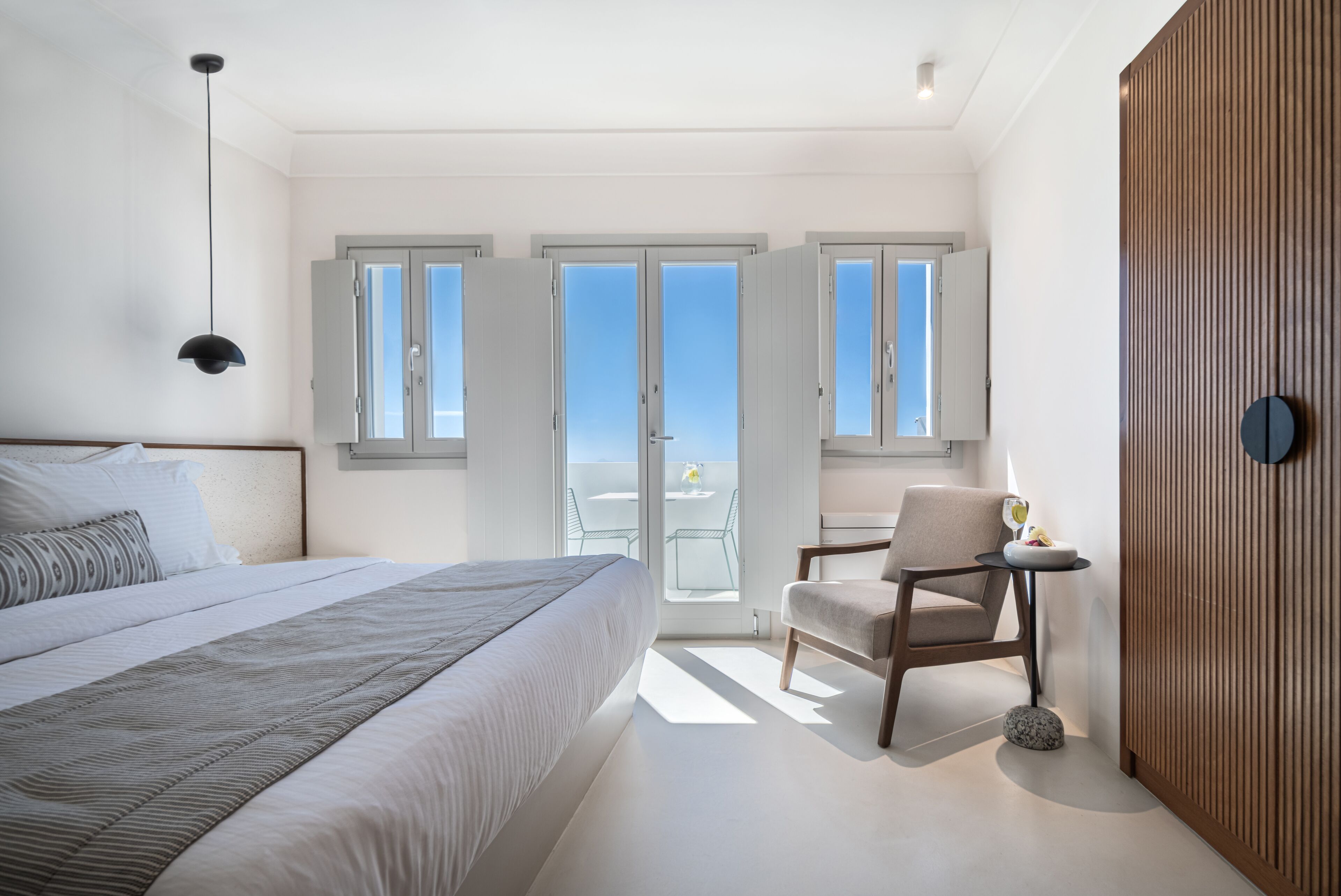 A modern bedroom with a large bed, a chair, and a small table. There are multiple windows providing natural light and a view of the outdoors.