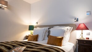 Una cama bien hecha con una manta y almohadas marrones, una mesita de noche con una lámpara y un termostato fijado a la pared.