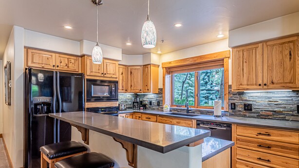 A modern kitchen with wooden cabinets, a large island with bar stools, a built-in microwave, and a stone backsplash. - Relaxed Comfort in a Prime Location – The Pines at Ore House! (Steamboat Springs)