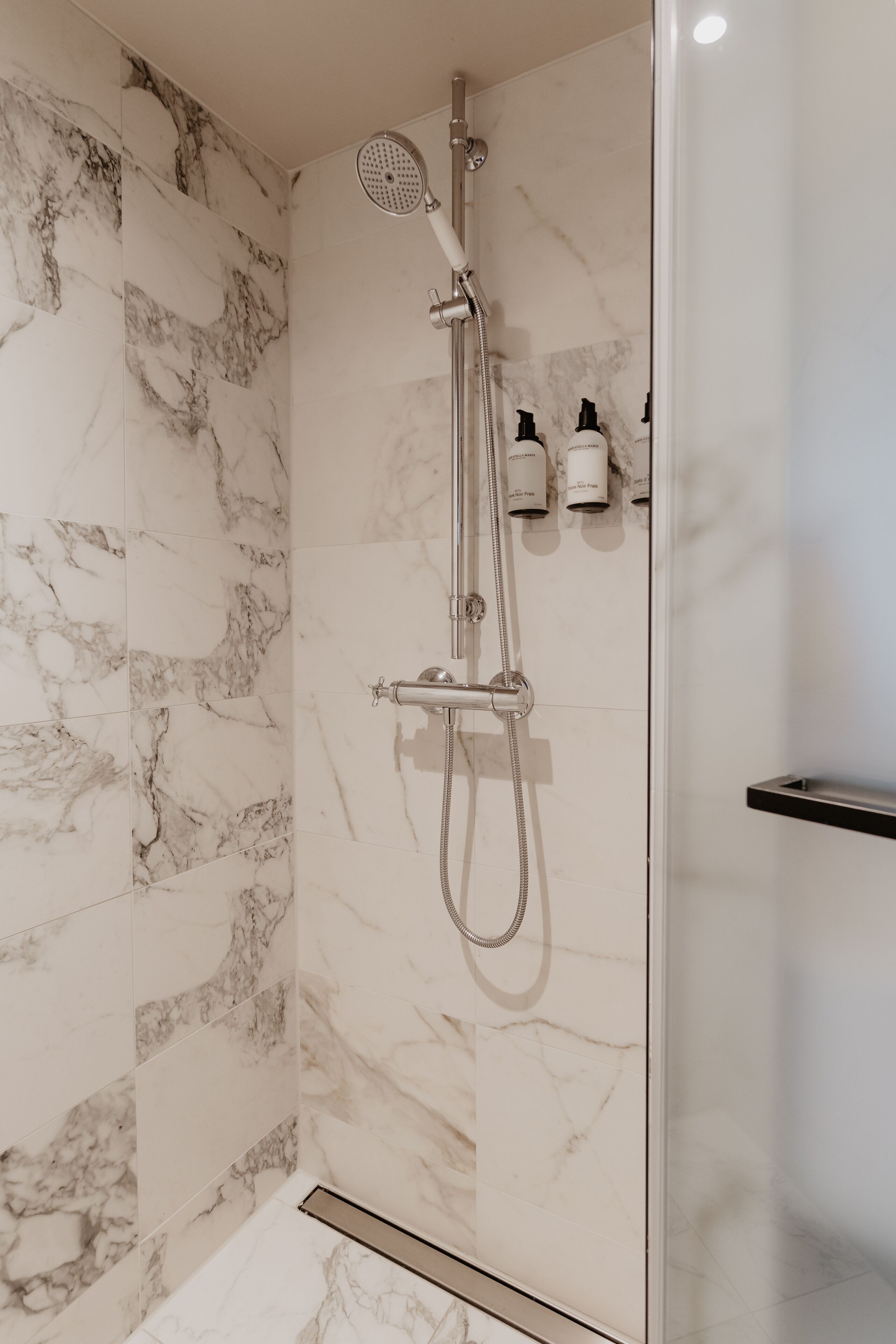 Een moderne douche met een handdouche, een kraan op de muur, marmeren tegels en een klein plankje met toiletartikelen.