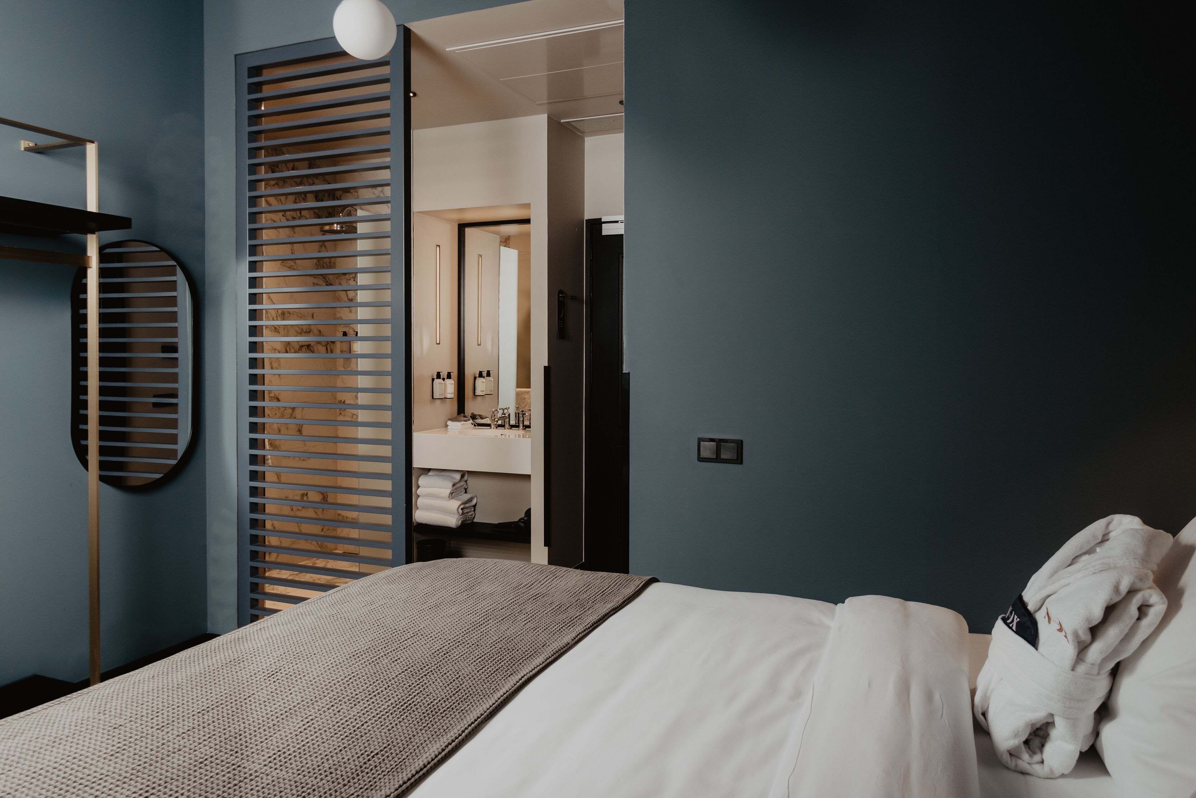 Una habitación de hotel moderna con una cama grande, un muro de listones de madera, un espejo redondo y un baño a la vista con lavamanos y perchero.