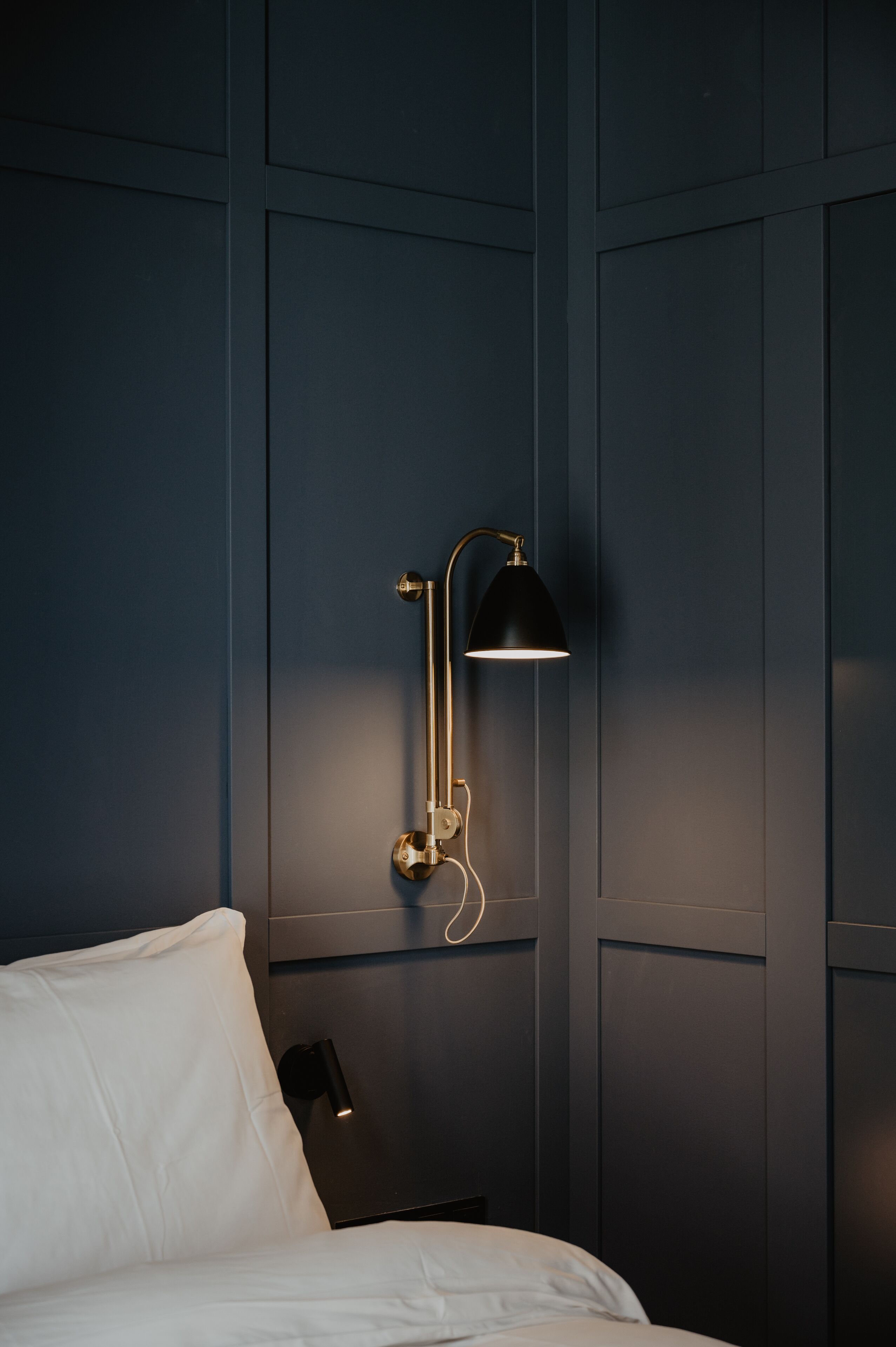 A dark blue wall with a black floor lamp and a white bed.