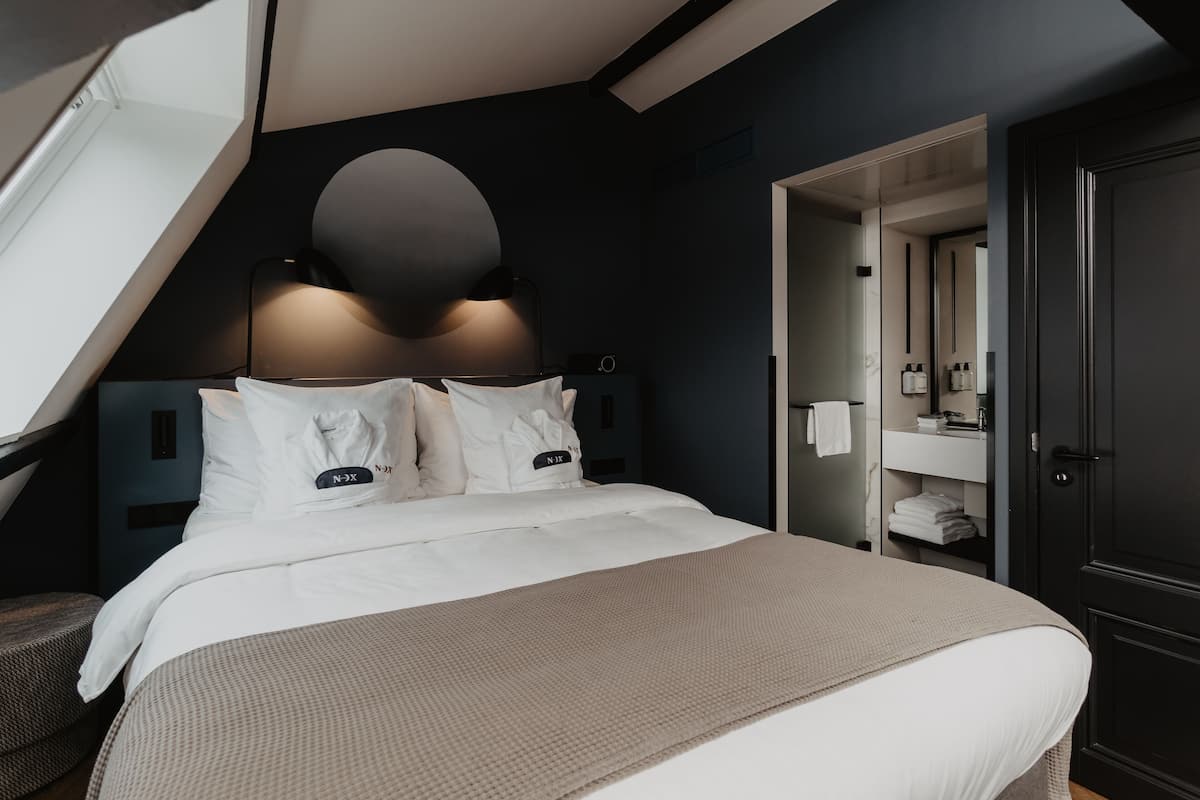 Ett modernt hotellrum med en stor säng, en rund vägglampa, en mörk garderob och ett badrum med synliga detaljer, inklusive en spegel och en handduksställning.