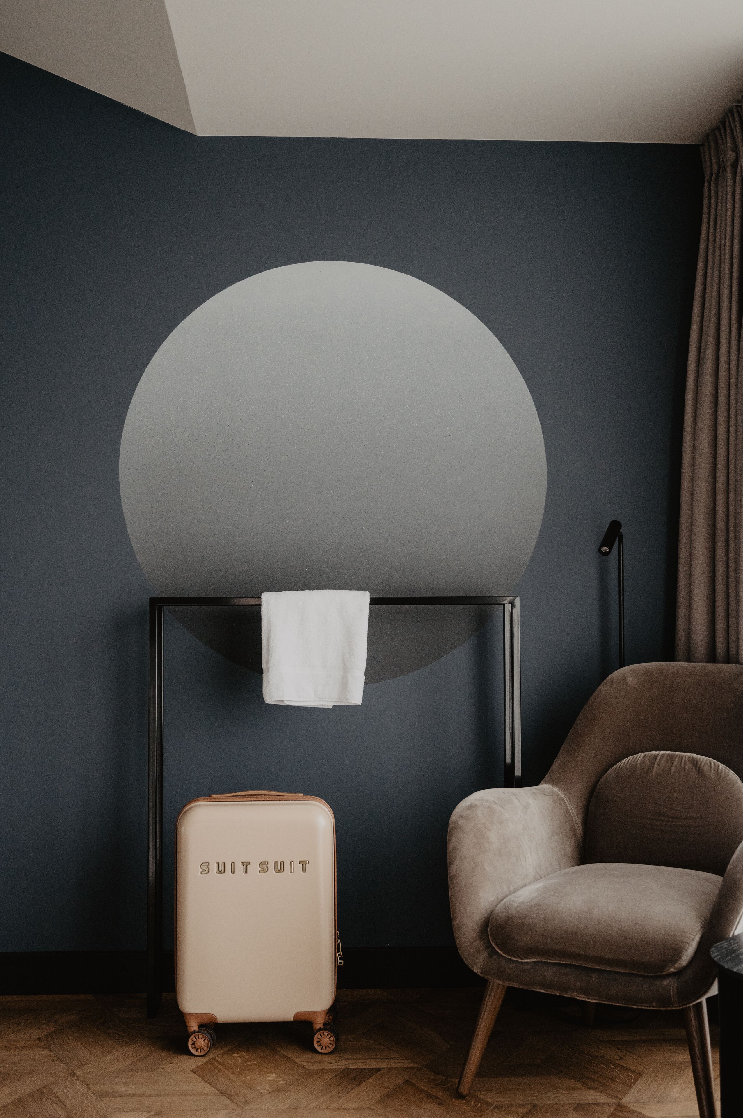 A room with a dark blue wall, a round light fixture, a beige suitcase, and a brown chair.