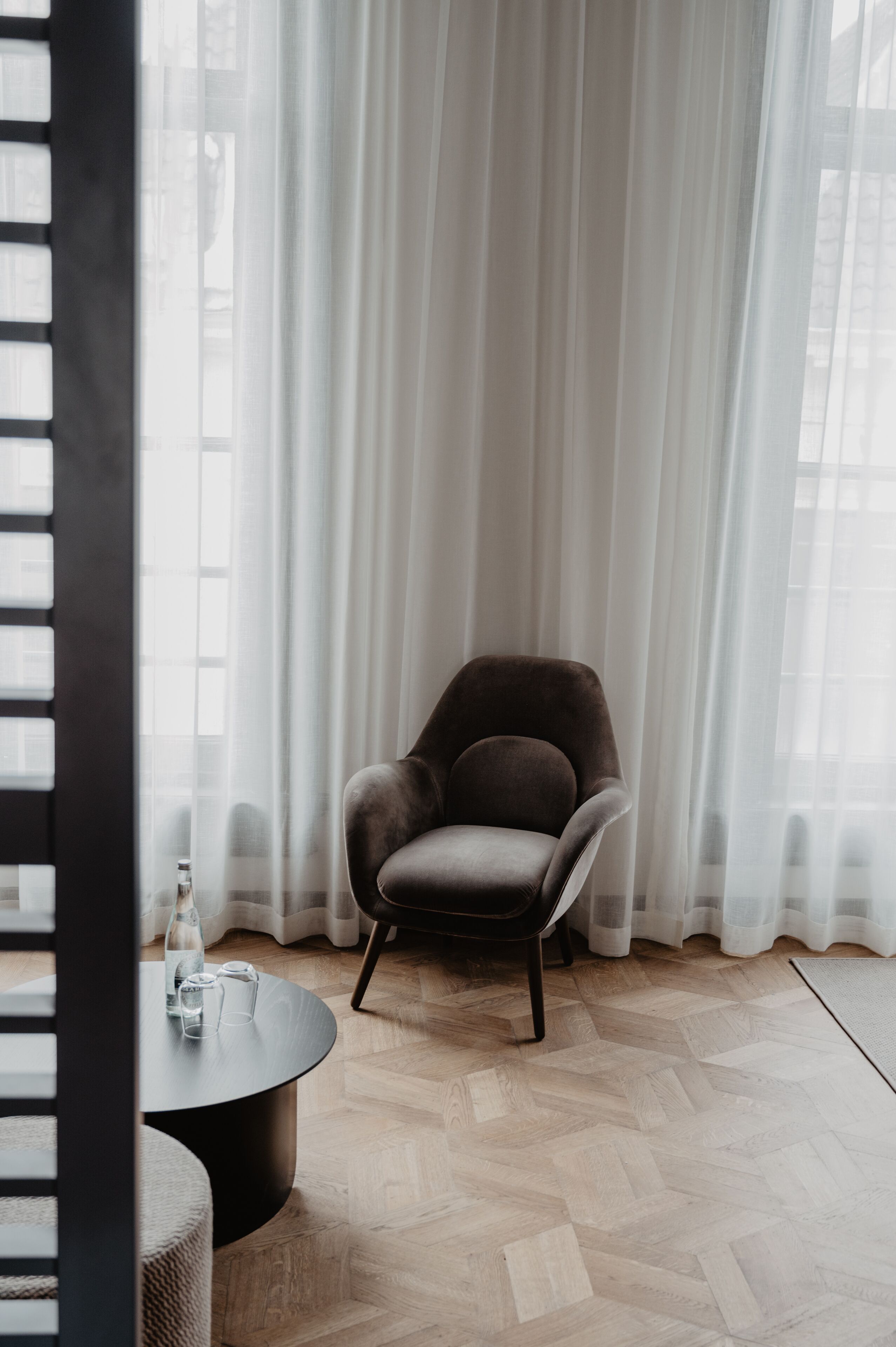A room with a dark brown armchair, a round black table with a bottle and glasses, and a herringbone wooden floor.
