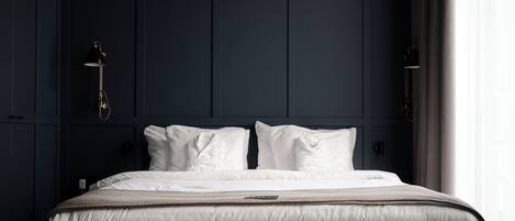 Una cama bien tendida con sábanas blancas y una colcha texturizada, frente a una pared oscura con una lámpara moderna.