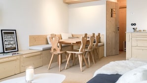 Une pièce minimaliste avec une table à manger en bois, des chaises, un canapé blanc, une petite table blanche avec une bougie et un livre, et un lit recouvert d’une couverture blanche et moelleuse.