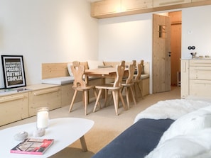 A minimalist room with a wooden dining table, chairs, a white sofa, a small white table with a candle and book, and a bed with a white furry cover. - Courchevel 1850: 3-bedroom apartment on slopes, parking (Courchevel 1850)