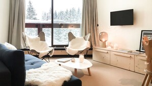 Un salon moderne comprenant un canapé, deux fauteuils, une table basse, un téléviseur et une vue sur des arbres enneigés, visible à travers de grandes fenêtres.