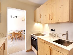 Une cuisine moderne avec des meubles en bois, un four encastré, un évier et un coin repas visible à travers une porte ouverte.