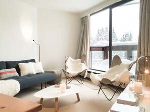 Un salon moderne avec un canapé bleu, des fauteuils blancs, une table basse blanche et une grande fenêtre donnant sur des arbres couverts de neige.