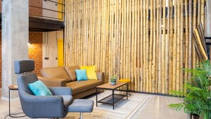 A modern lounge area with a bamboo wall, a brown sofa with blue and yellow cushions, a grey armchair, a small round table with a plant, and a potted plant to the side. - Art Loft 06 'Travelling Light' in Valencia (València)