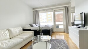 A modern hotel room with a white sofa, a glass coffee table, a bed with grey bedding, a TV on a dresser, and a view of buildings through the window. - City Home Finland Laukontori (Tampere)