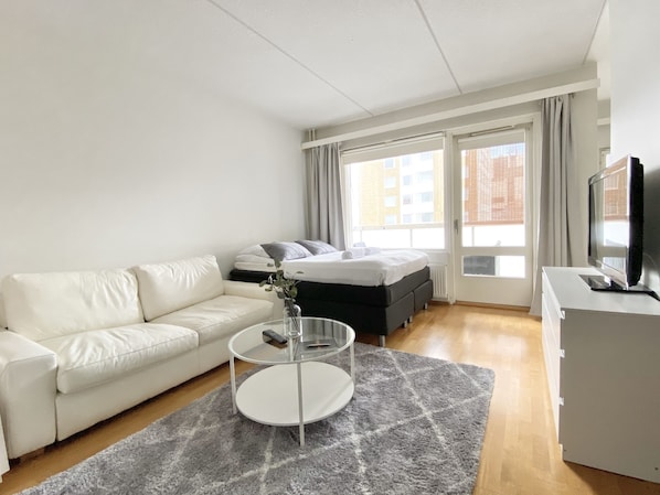 A modern hotel room with a white sofa, a bed with grey bedding, a glass coffee table, and a television on a white cabinet.