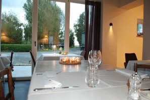 A dining table set with candles, a wine glass, and salt and pepper shakers, with a view of a garden and street outside.