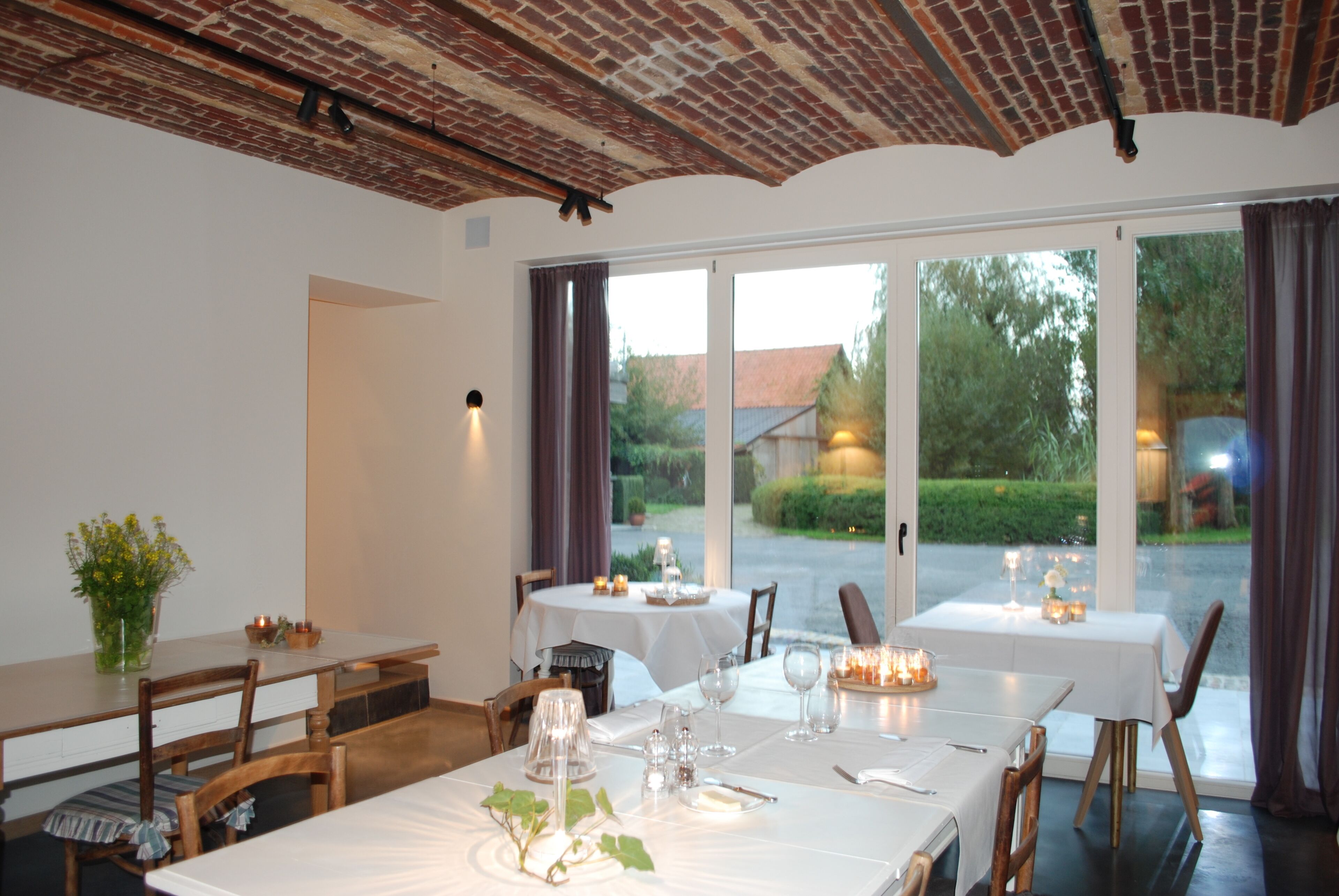 A restaurant with two tables set for dining, large windows, and a brick ceiling.