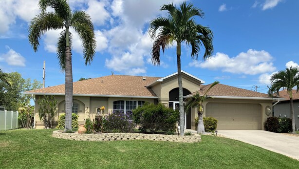 A single-story house with a beige exterior, a tiled roof, and a front yard with palm trees and a well-maintained lawn. - Cape Escape SW Florida (Cape Coral)