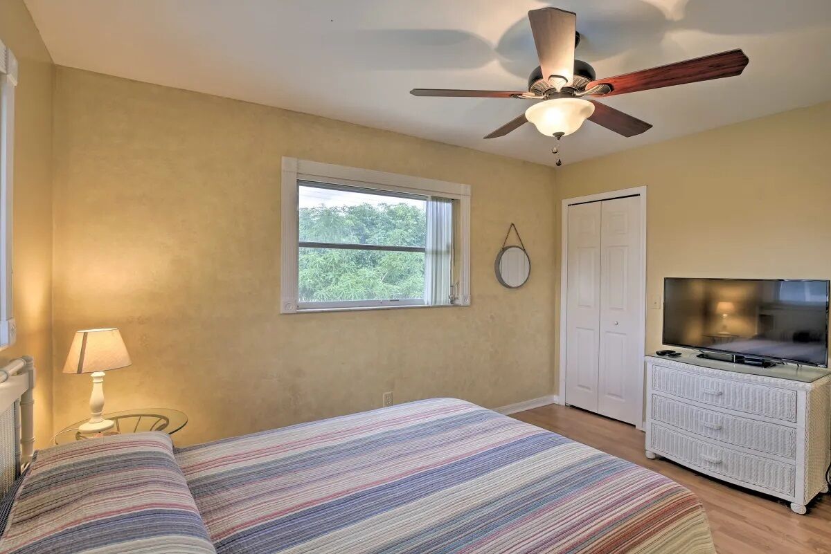 A bedroom with a bed, a ceiling fan, a TV, a dresser, a window, and a door.
