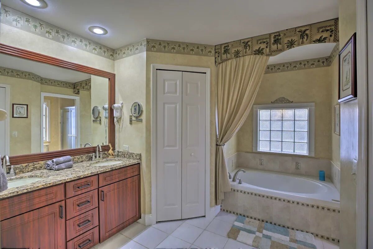 A bathroom with a large mirror, a sink with a granite countertop, a vanity with drawers, a door, and a bathtub with a window.