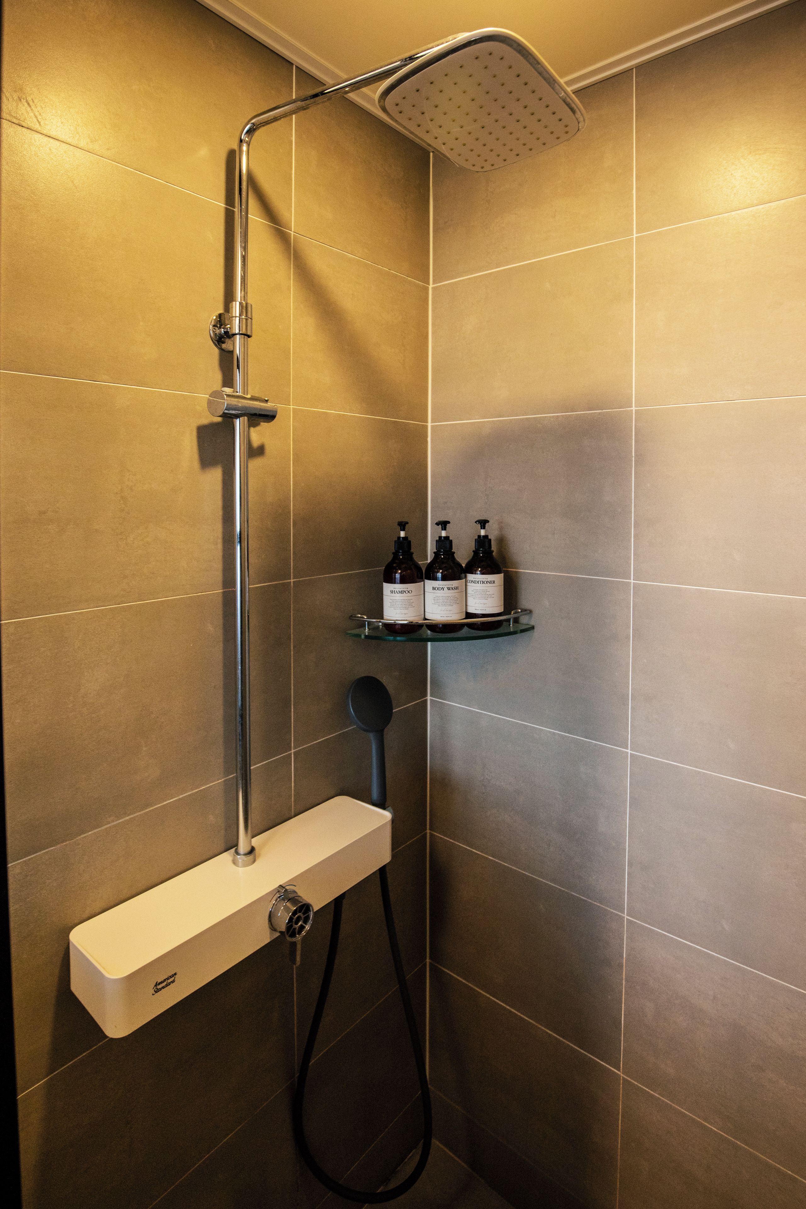 Zona de ducha con cabezal de ducha manual, un accesorio de ducha de mano y un estante con tres botellas de champú, gel de baño y acondicionador.