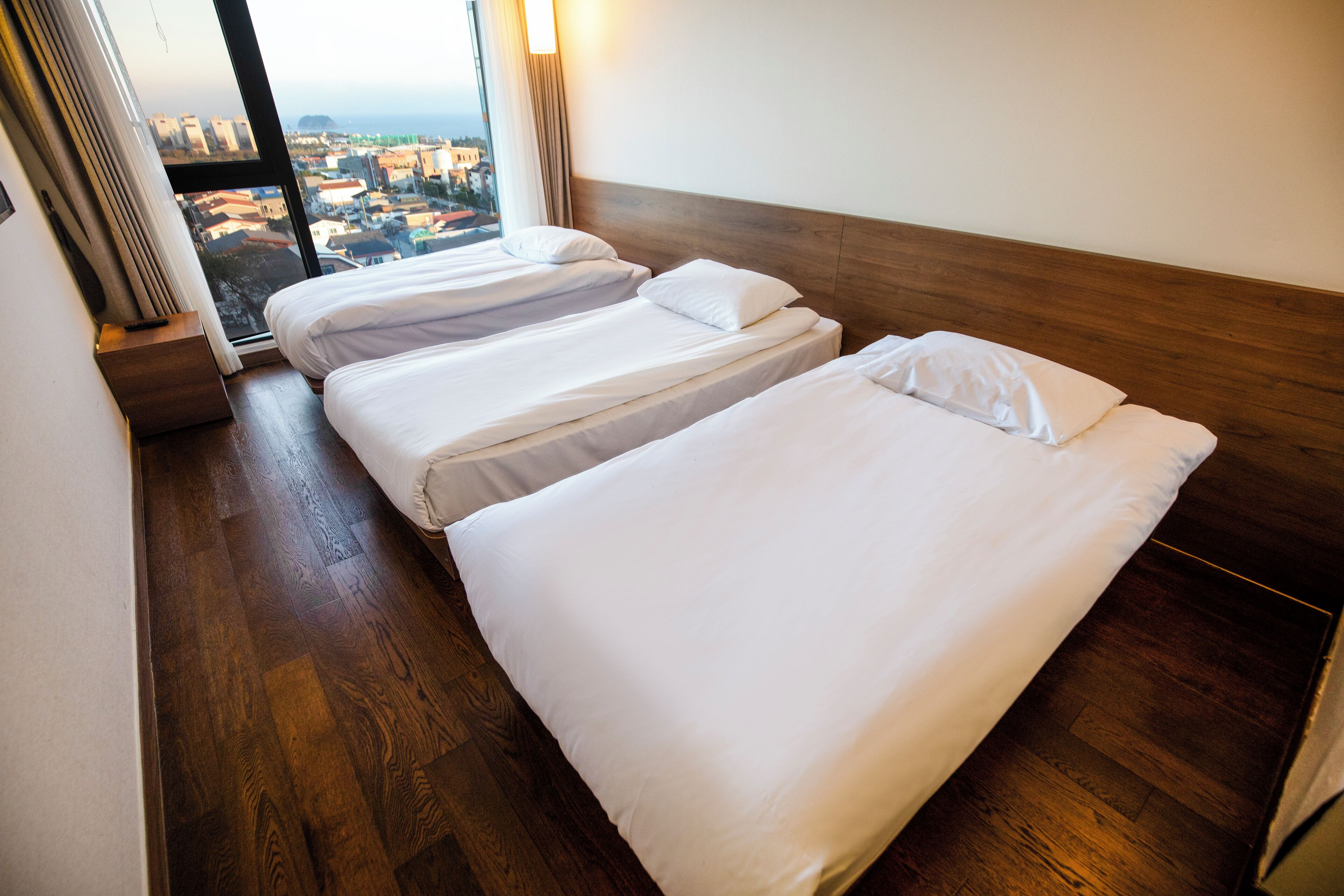 A hotel room with two single beds, wooden flooring, a large window with a city view, and a wooden headboard.