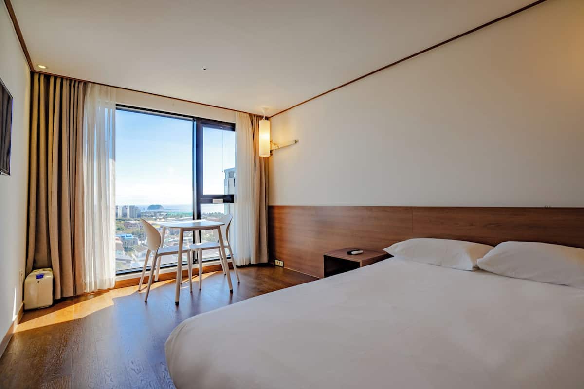 Ein Hotelzimmer mit einem großen Bett, einem Schreibtisch mit Stuhl, Blick auf die Stadt durch das Fenster und einem Nachttisch mit Lampe.