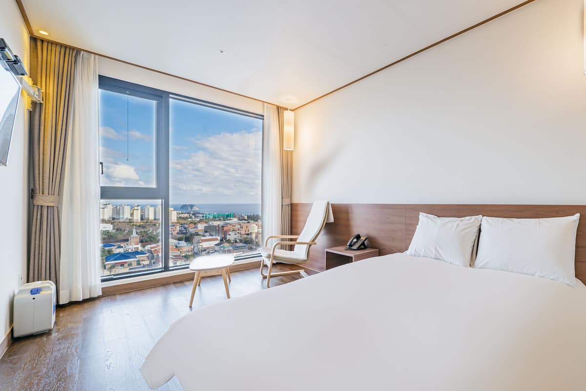 Ein Hotelzimmer mit einem großen Bett, einem Sessel, einem Schreibtisch und Blick auf die Stadt durch ein großes Fenster.