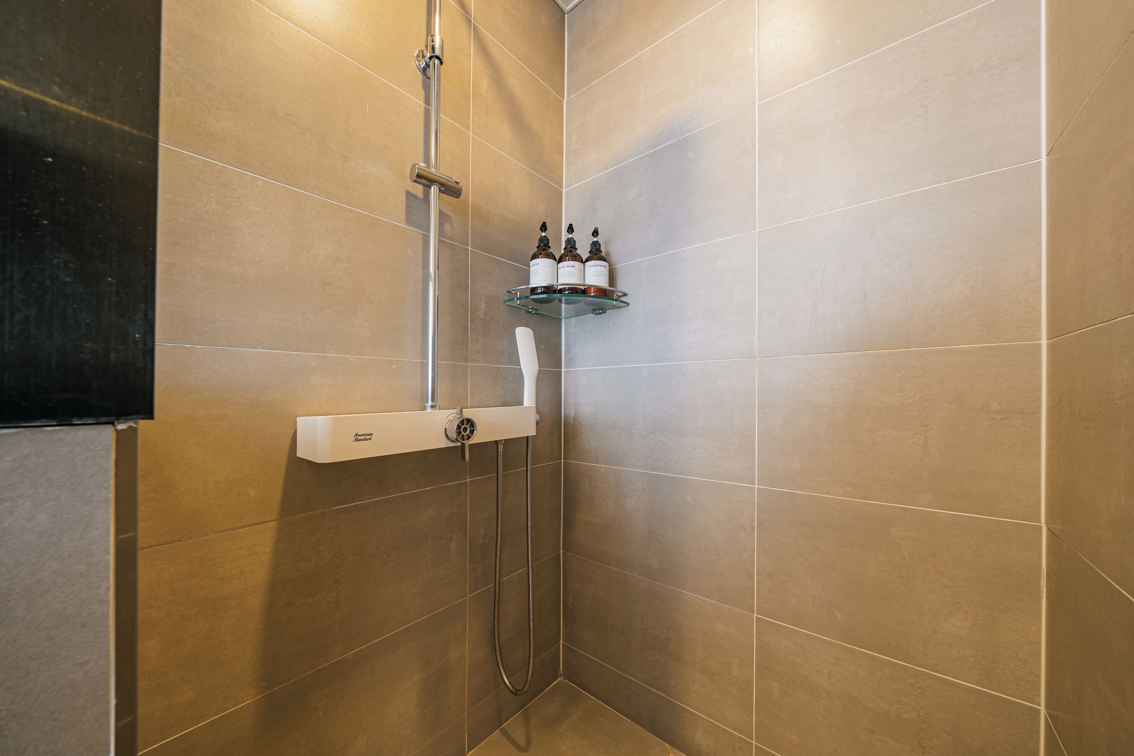 A shower area with a handheld showerhead, a wall-mounted control panel, and a glass shelf with toiletries.
