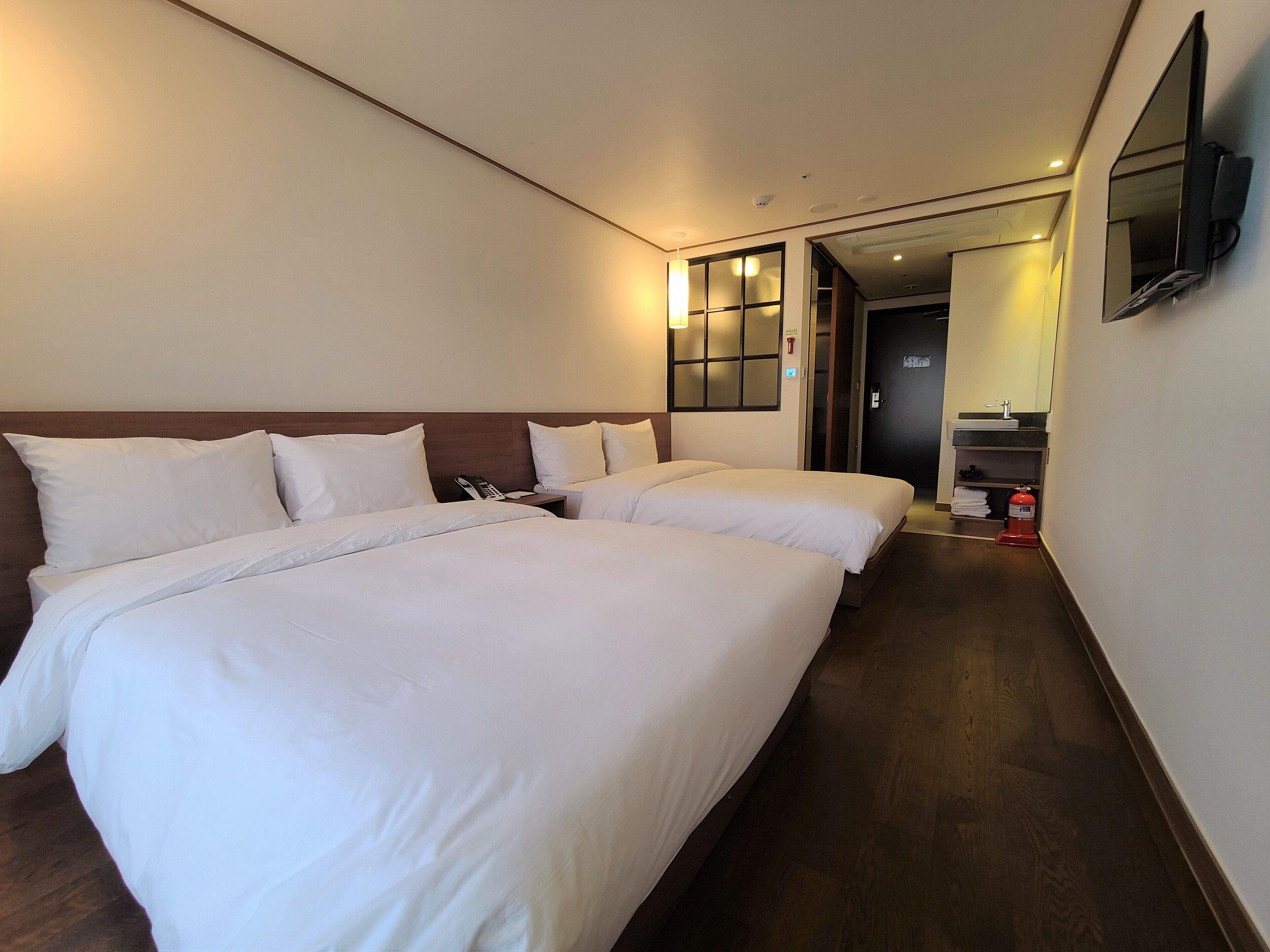 A hotel room with two beds, a wooden headboard, a flat-screen TV mounted on the wall, and a small bathroom with a sink and mirror.