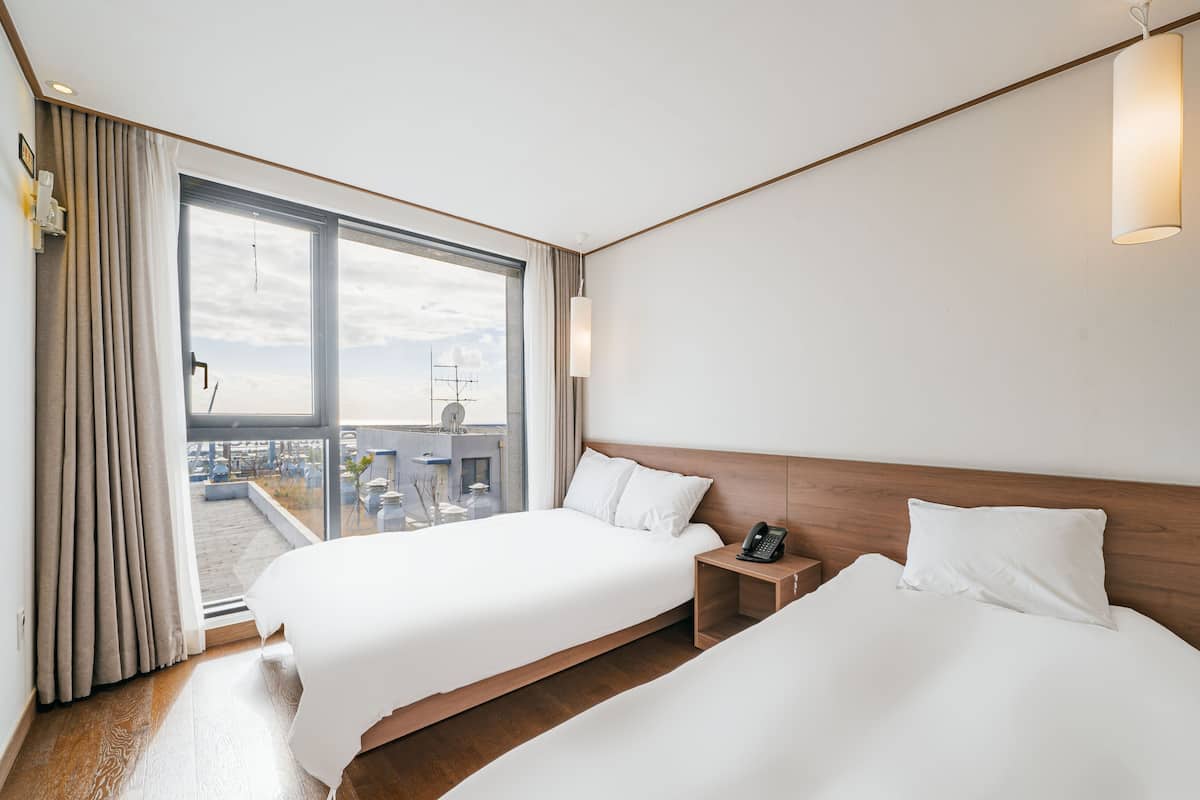 Ein Hotelzimmer mit zwei Betten, einem großen Fenster mit Vorhängen, einem Nachttisch mit Telefon und Blick auf die Stadt.