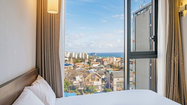 A hotel room with a large window, a bed with white linens, and curtains. - Tamara Jeju (Seogwipo)