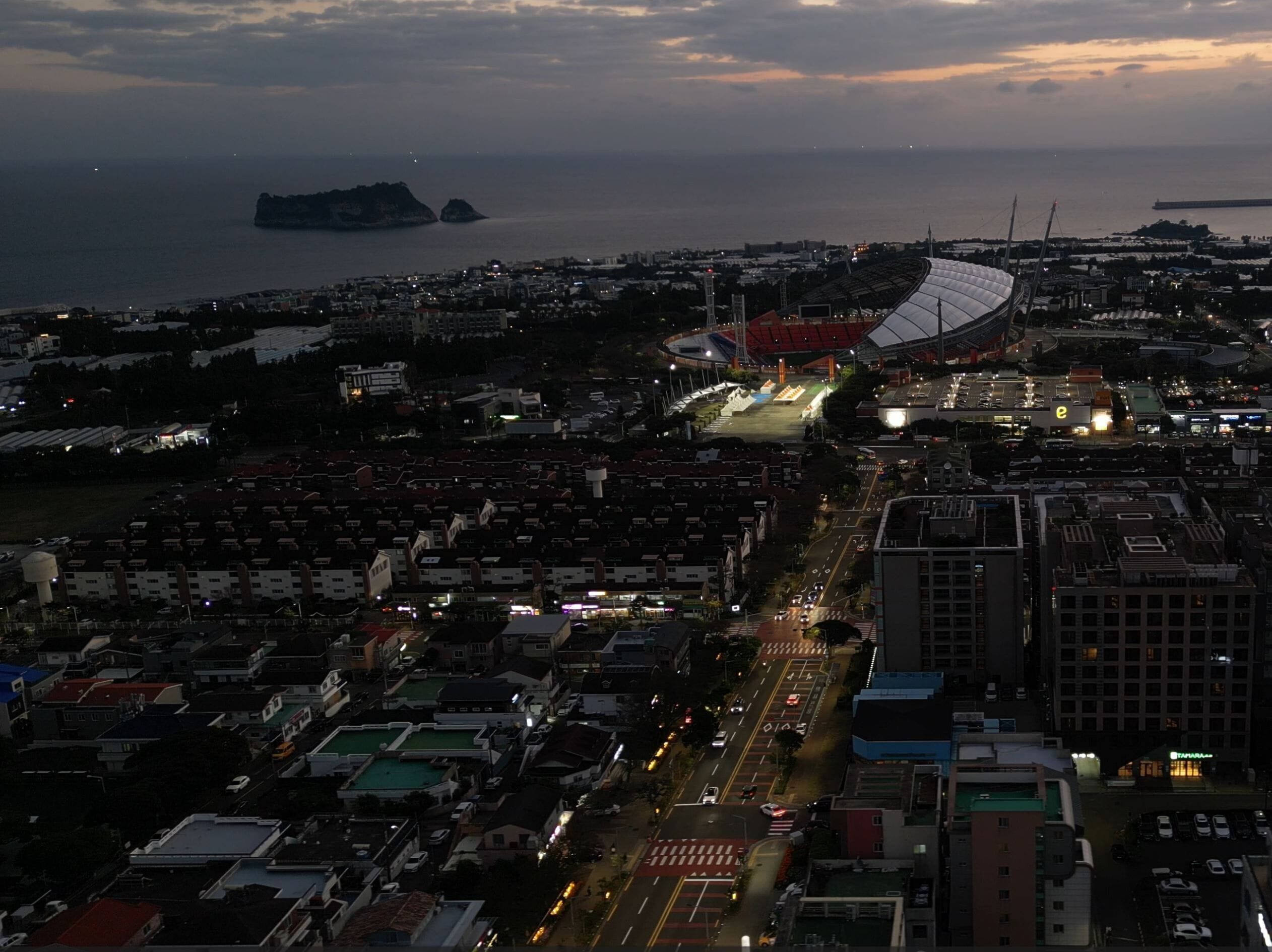 A stadium with a distinctive roof structure, surrounded by urban buildings and a coastline.