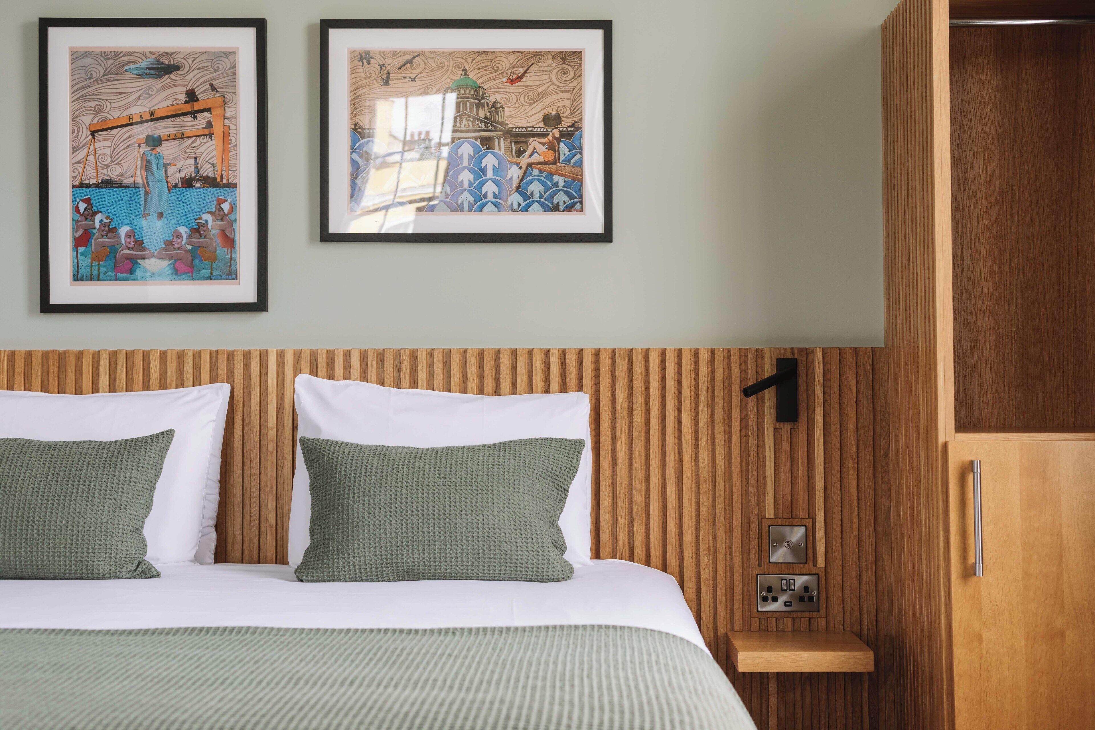 A hotel room with a wooden headboard, two green pillows, a bed with white bedding, and two framed artworks on the wall.