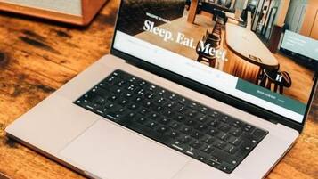 Una computadora portátil sobre una mesa de madera mostrando una página web con el texto 'Sleep, Eat, Meet'.