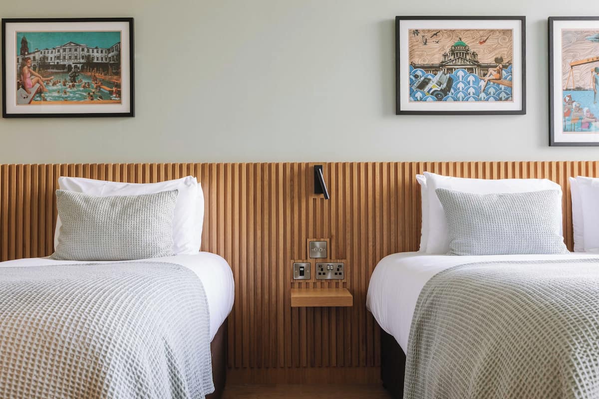 A hotel room with two beds, wooden headboard, a small bedside table with a lamp, and three framed artworks on the wall.