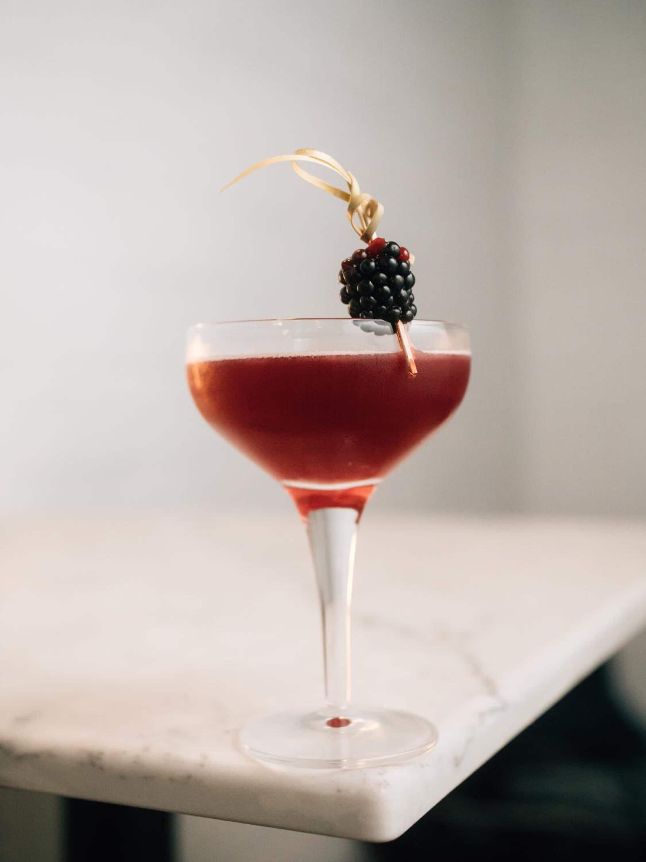 A martini glass with a red drink, a blackberry garnish, and a gold ribbon on a marble surface.