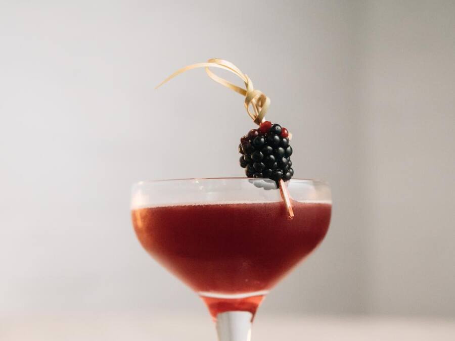 A martini glass with a red drink, a blackberry garnish, and a gold ribbon on a marble surface.